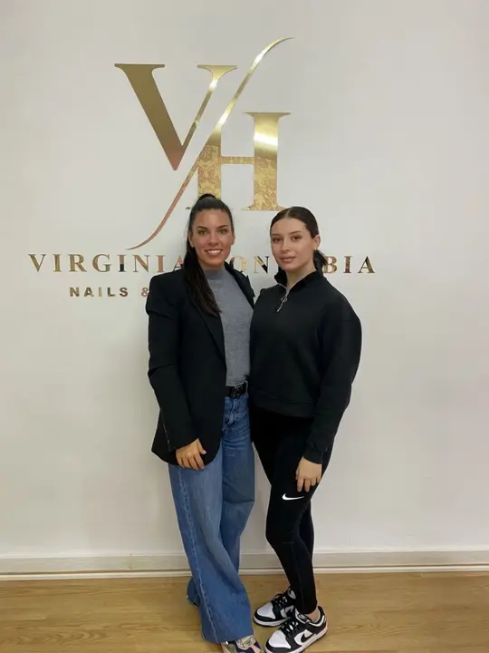 Virginia Honrubia junto a su alumna Paula, mirando al frente y sonriendo, frente al logo de Virginia Honrubia Nails &amp; Studio Academy, reflejando formaci&oacute;n profesional, trabajo en equipo y excelencia en el sector de la manicura.
