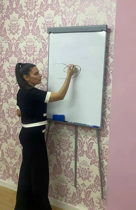 Virginia Honrubia de pie escribiendo en una pizarra durante una clase de manicura profesional, mostrando su experiencia como instructora y su compromiso con la formaci&oacute;n de futuras profesionales del sector de las u&ntilde;as.