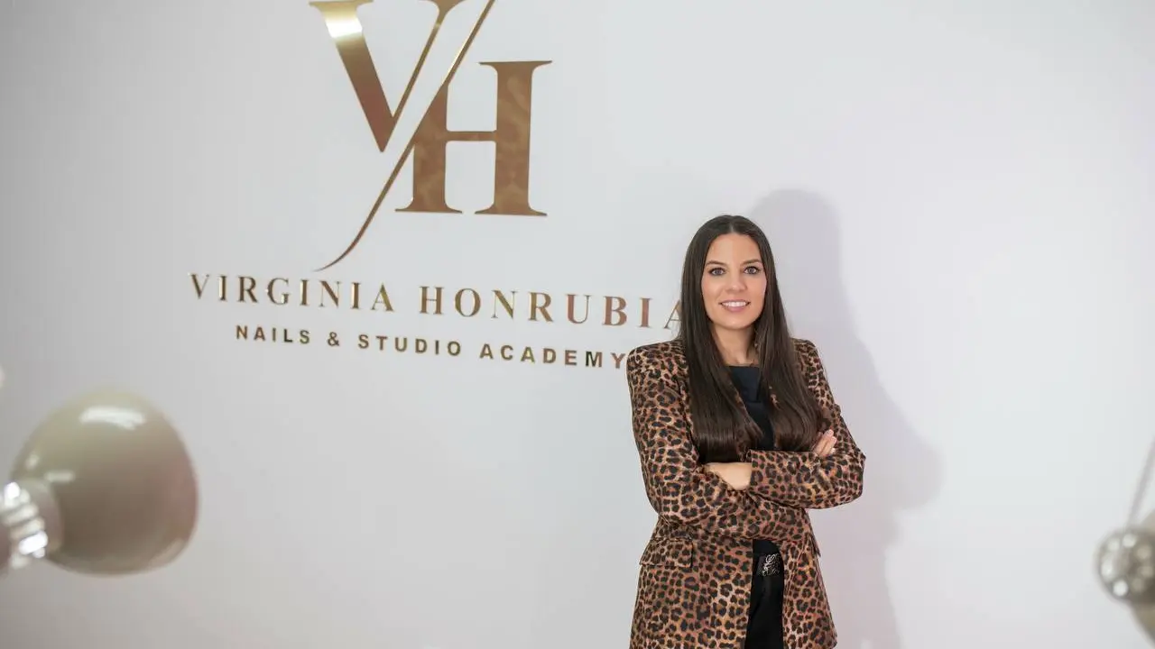 En la imagen aparece Virginia dentro de su sal&oacute;n de u&ntilde;as, posando con seguridad y profesionalismo mientras detr&aacute;s de ella se aprecia claramente el logo oficial del negocio en la pared. El entorno refleja un espacio moderno, limpio y bien iluminado, reforzando la identidad de marca y la imagen profesional del sal&oacute;n. La presencia del logotipo en el fondo aporta reconocimiento visual y transmite confianza a las clientas.