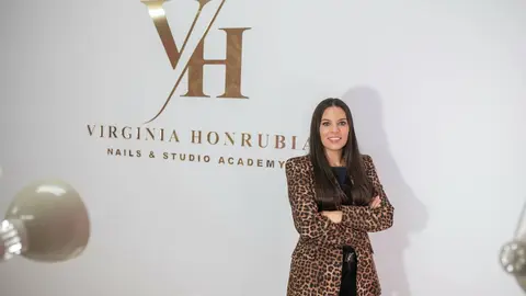 En la imagen aparece Virginia dentro de su sal&oacute;n de u&ntilde;as, posando con seguridad y profesionalismo mientras detr&aacute;s de ella se aprecia claramente el logo oficial del negocio en la pared. El entorno refleja un espacio moderno, limpio y bien iluminado, reforzando la identidad de marca y la imagen profesional del sal&oacute;n. La presencia del logotipo en el fondo aporta reconocimiento visual y transmite confianza a las clientas.