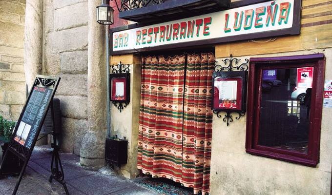 Vista exterior de la fachada del histórico Bar Ludeña, situado en el casco antiguo de Toledo, con su característico letrero y cortina y un entorno de calles estrechas típicas del centro histórico, reflejando el ambiente tradicional de la ciudad.